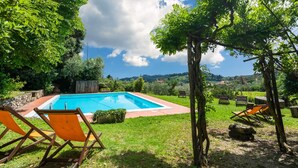 A rectangular swimming pool surrounded by grass, with two wooden chairs and a pergola in the foreground. - Italian Stay: Bacchereto Wine Country (Carmignano)