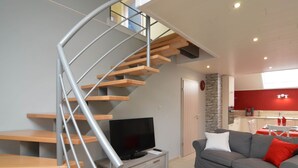 A modern living room with a spiral staircase, a flat-screen TV on a wooden console, a grey sofa with red cushions, and a kitchen in the background. - A Holiday Home Ideal for Families and for Discovering the Ardennes (Waimes)