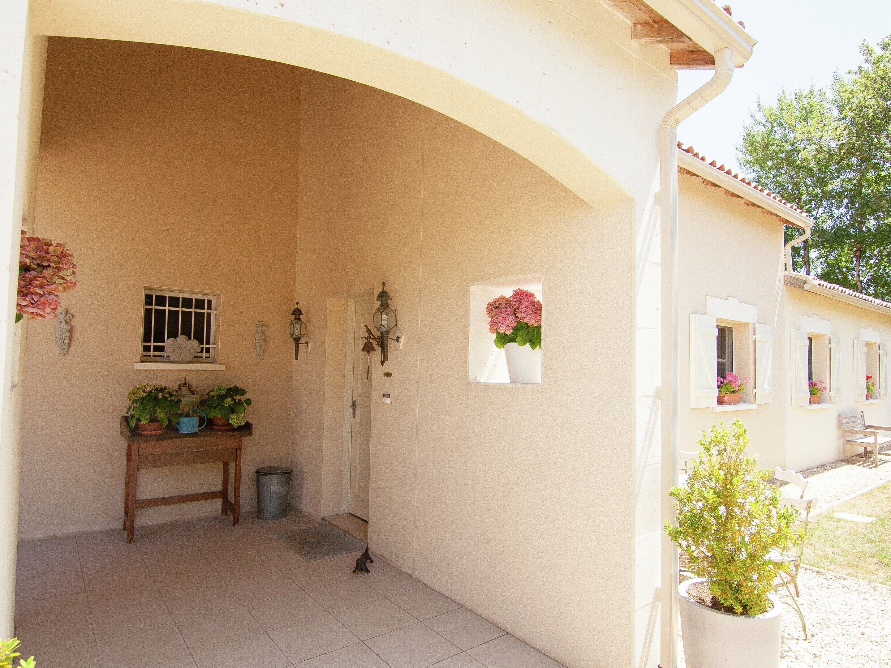 A covered porch with a tiled floor, a small table with potted plants, a door, and a window with a view of the outside.