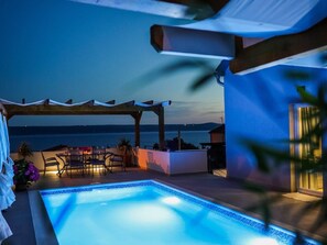 A pool area with a pergola, seating, and a view of the sea at dusk.