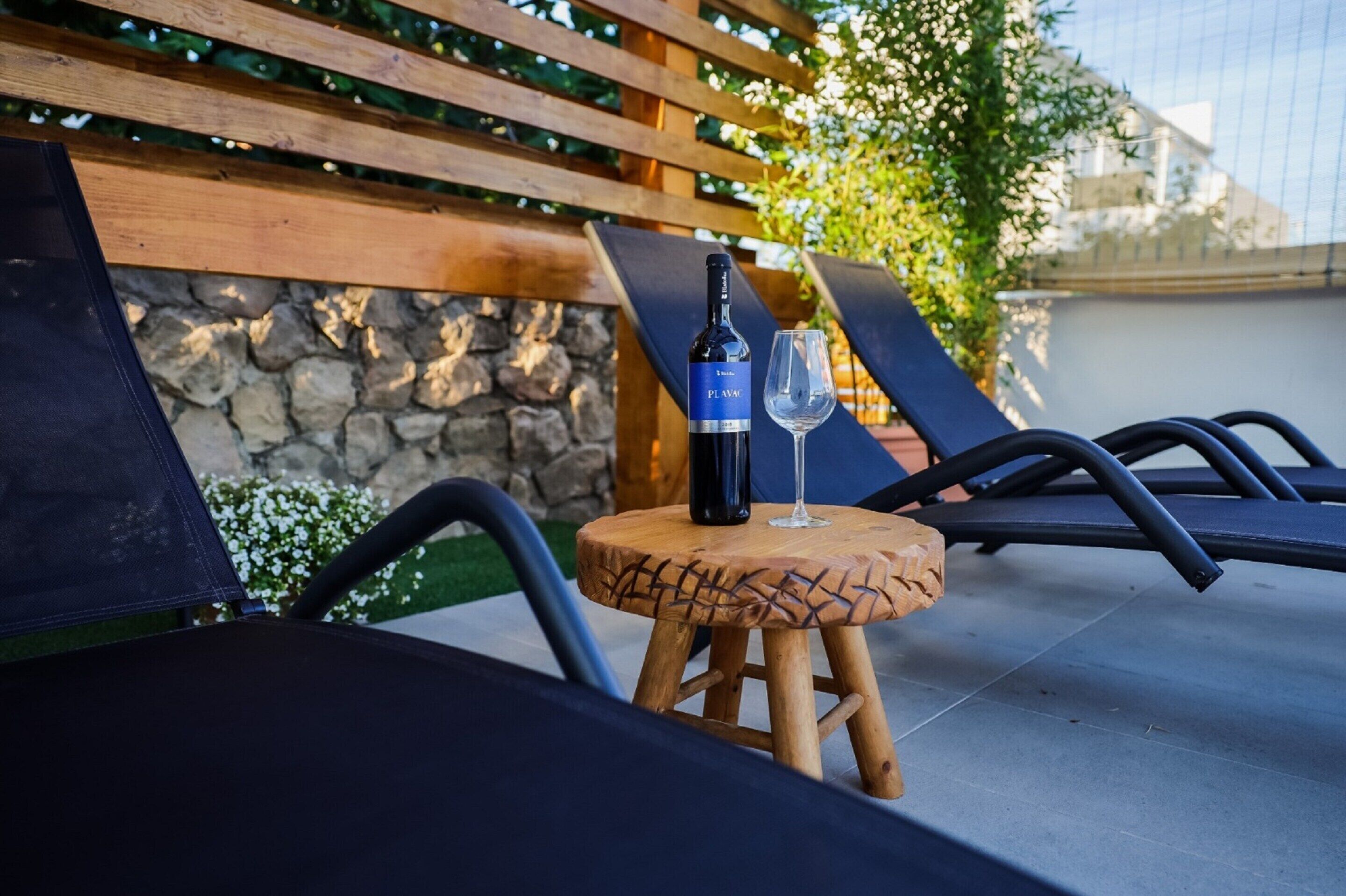 Une bouteille de vin Plasmic et un verre posés sur un tabouret en bois, dans un espace extérieur aménagé avec des fauteuils et un mur en pierre.