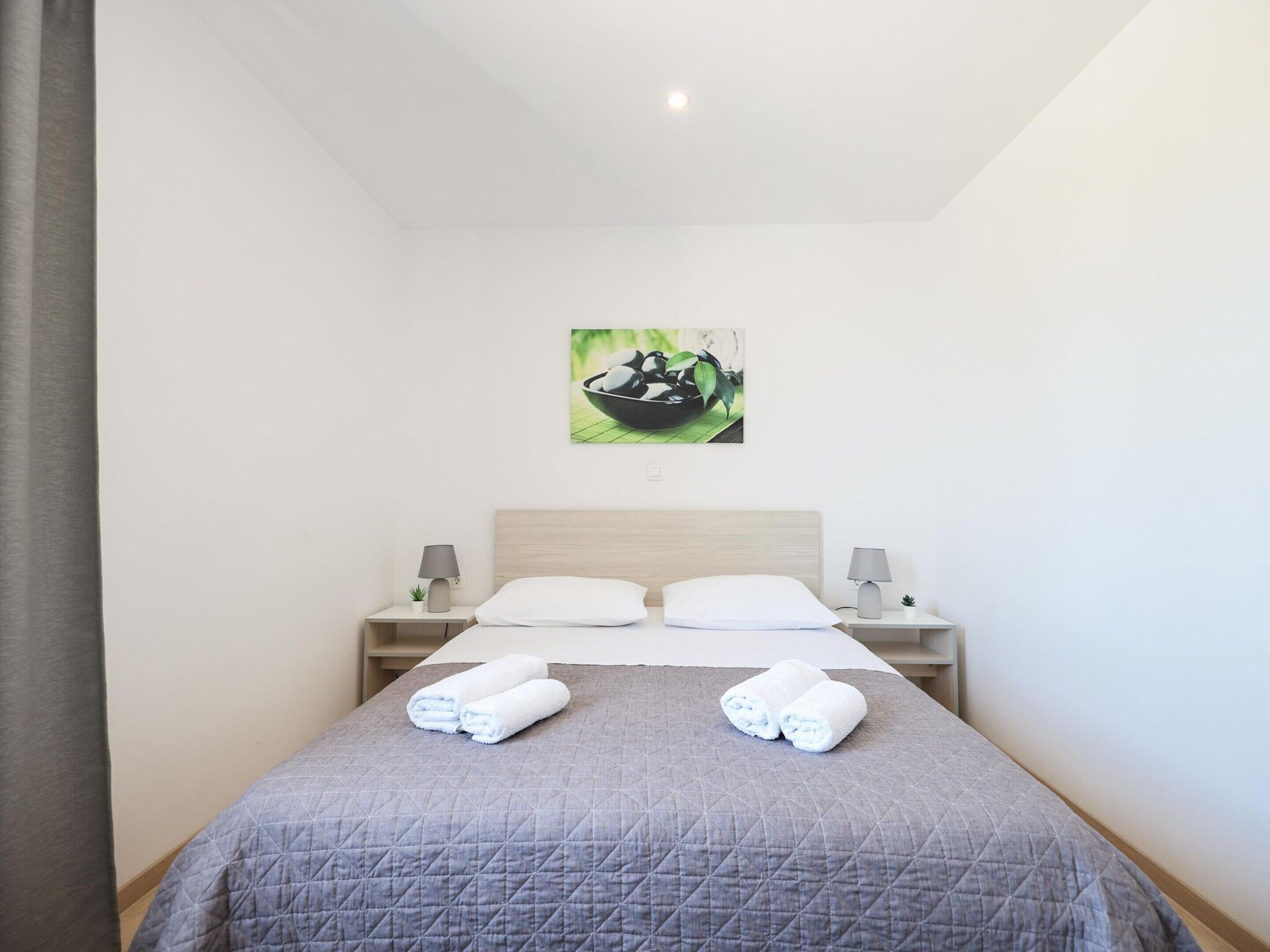 Un lit bien fait, recouvert d’une couverture grise, avec deux oreillers blancs et deux serviettes enroulées posées sur le lit. Une petite table avec une lampe et une plante se trouve à côté du lit. Un tableau représentant un bol rempli de plantes est accroché au mur, au-dessus du lit.