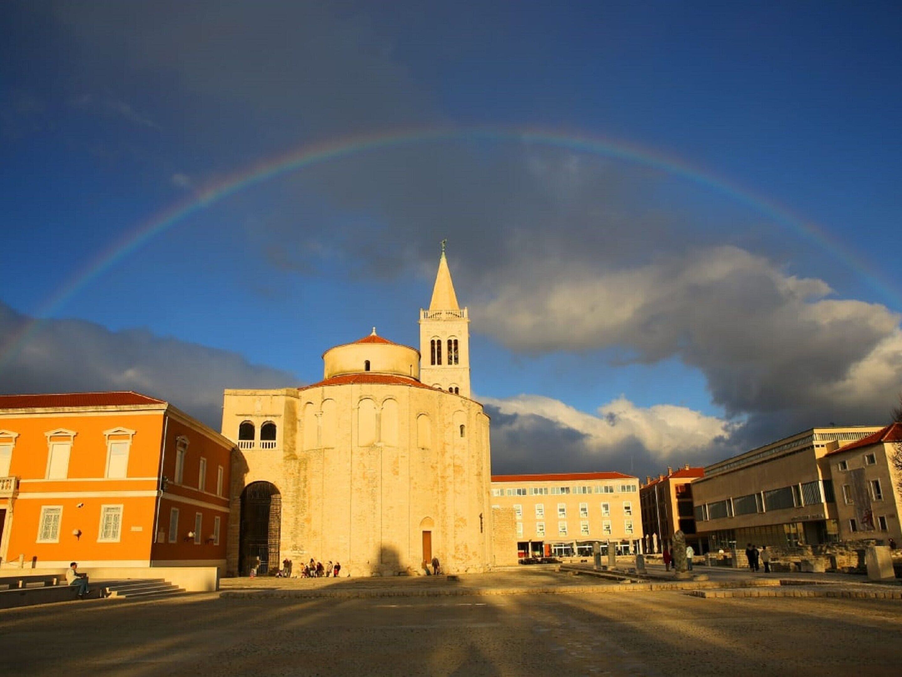 Ein historisches Gebäude mit einem markanten Turm und einer Kuppel, umgeben von modernen Bauten, unter wolkenlosem Himmel mit einem Regenbogen.