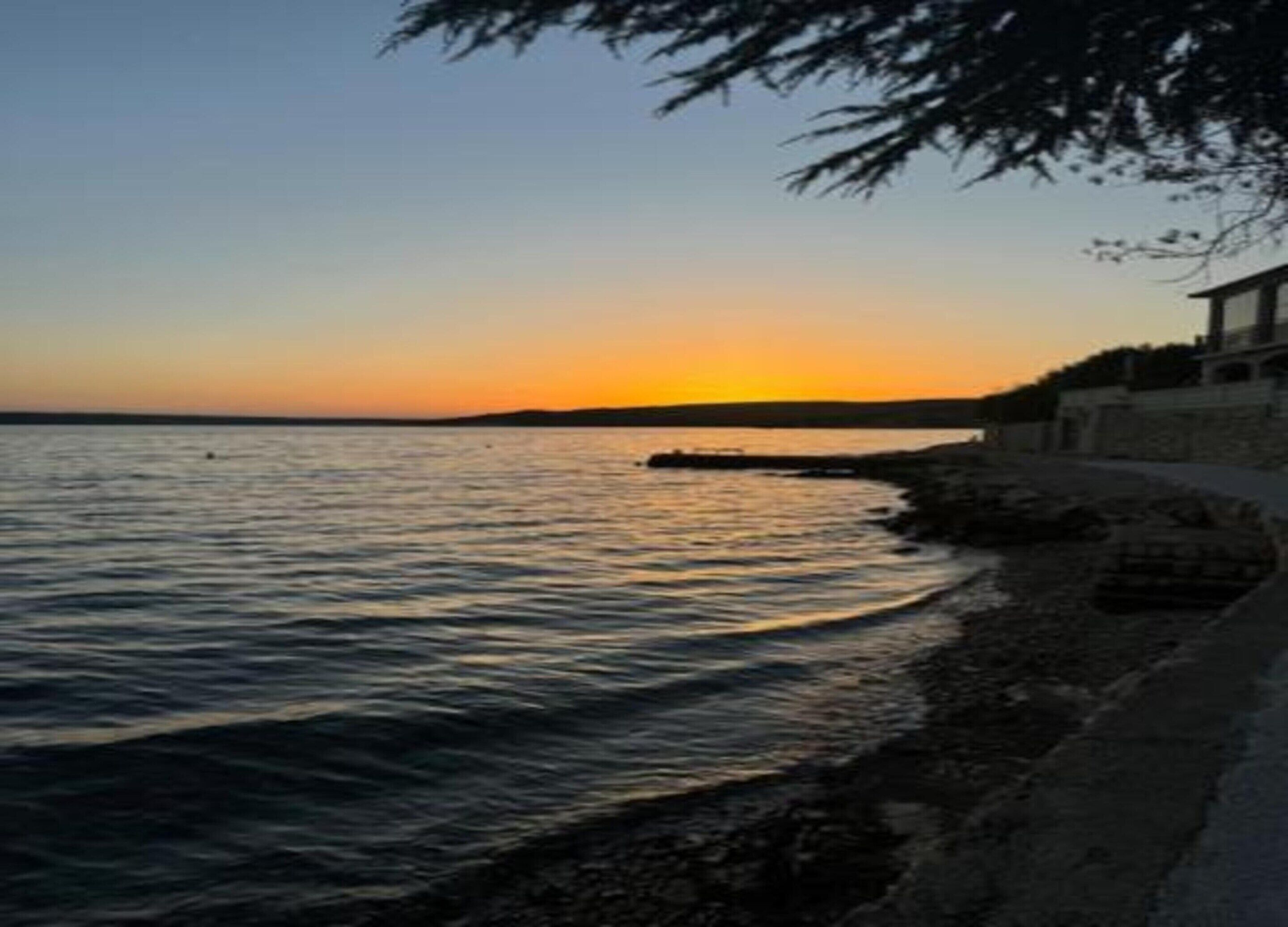 A coastal scene with a sunset over the water, a stone wall, and a building with a balcony.