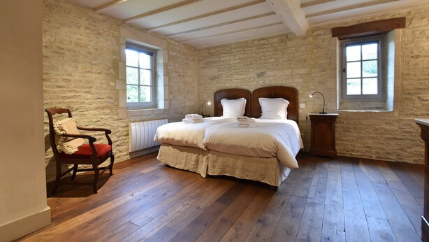 Ein Schlafzimmer mit Steinwand, Holzboden, einem Bett mit weißen Leinen, einem Holzstuhl und zwei Fenstern.
