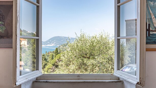 Une fenêtre ouverte donnant sur un paysage côtier, avec des arbres et une montagne lointaine.