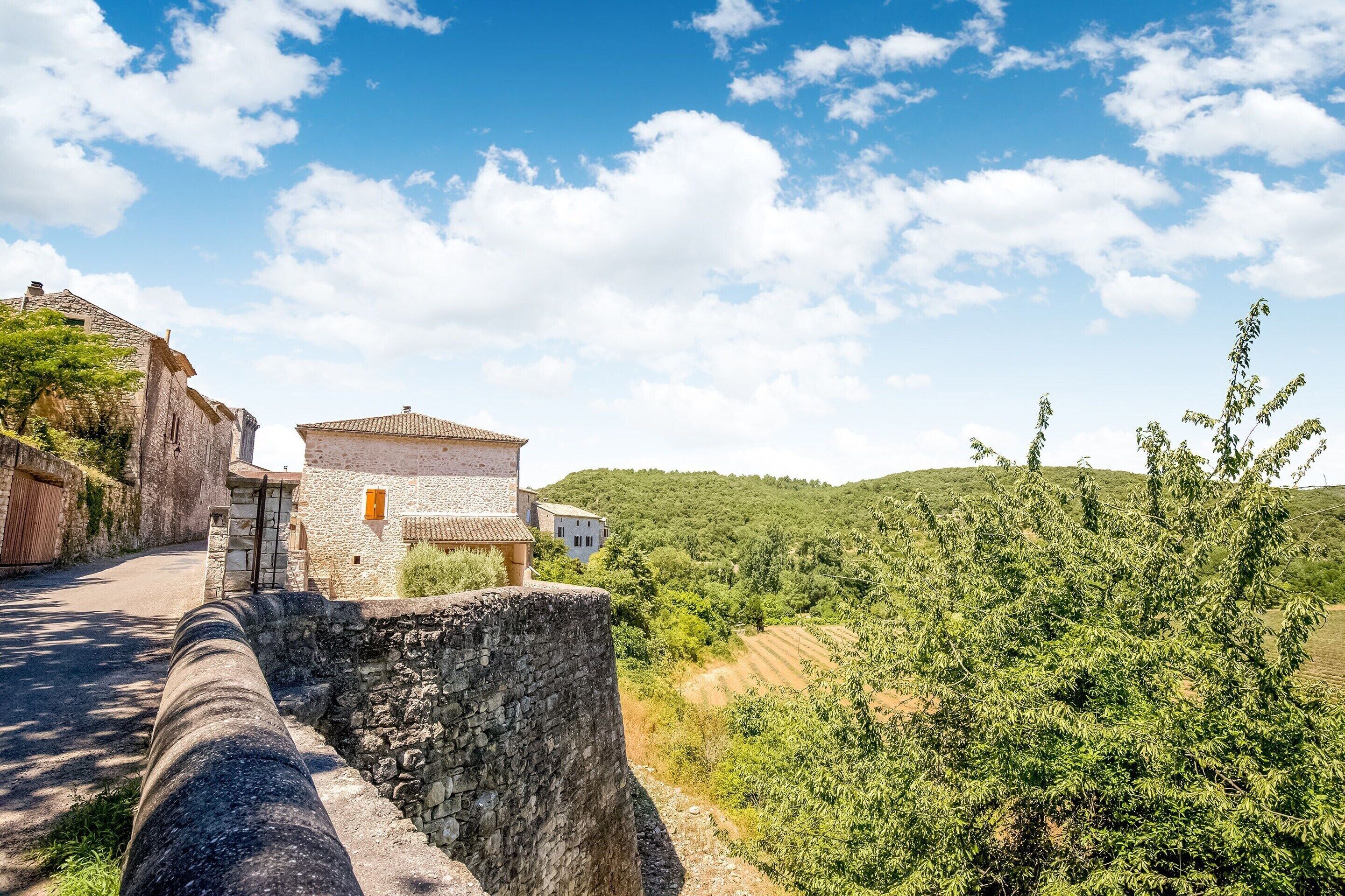 A stone pathway leading to a rustic building with a tiled roof, surrounded by lush greenery and hills.