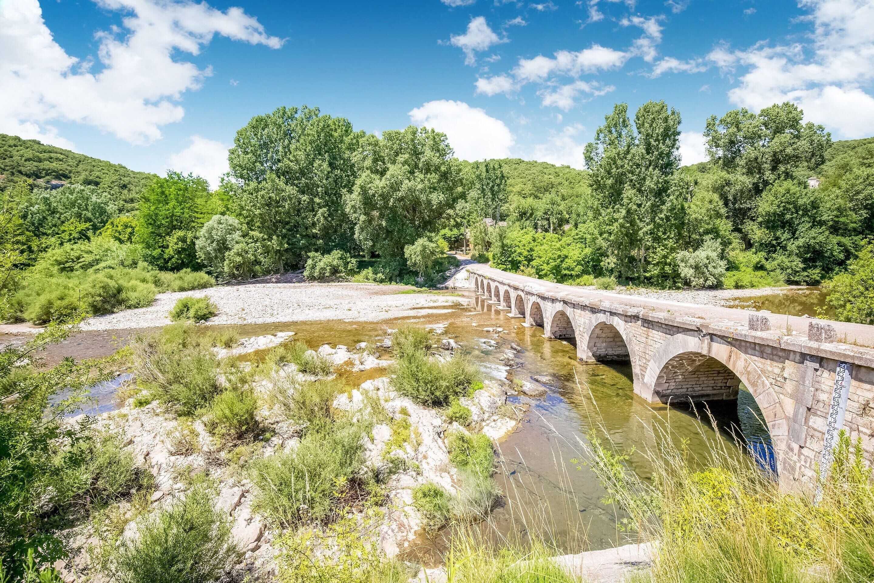 A stone bridge over a river with surrounding greenery and hills.