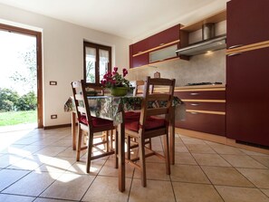 Uma área de refeições com uma mesa posta para quatro pessoas, cercada por cadeiras de madeira com almofadas vermelhas. Um vaso verde com flores está sobre a mesa. O ambiente possui armários de madeira e uma janela com vista para o verde lá fora.