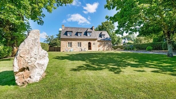 Una casa de piedra con una gran roca al frente, rodeada de césped cuidado y árboles.
