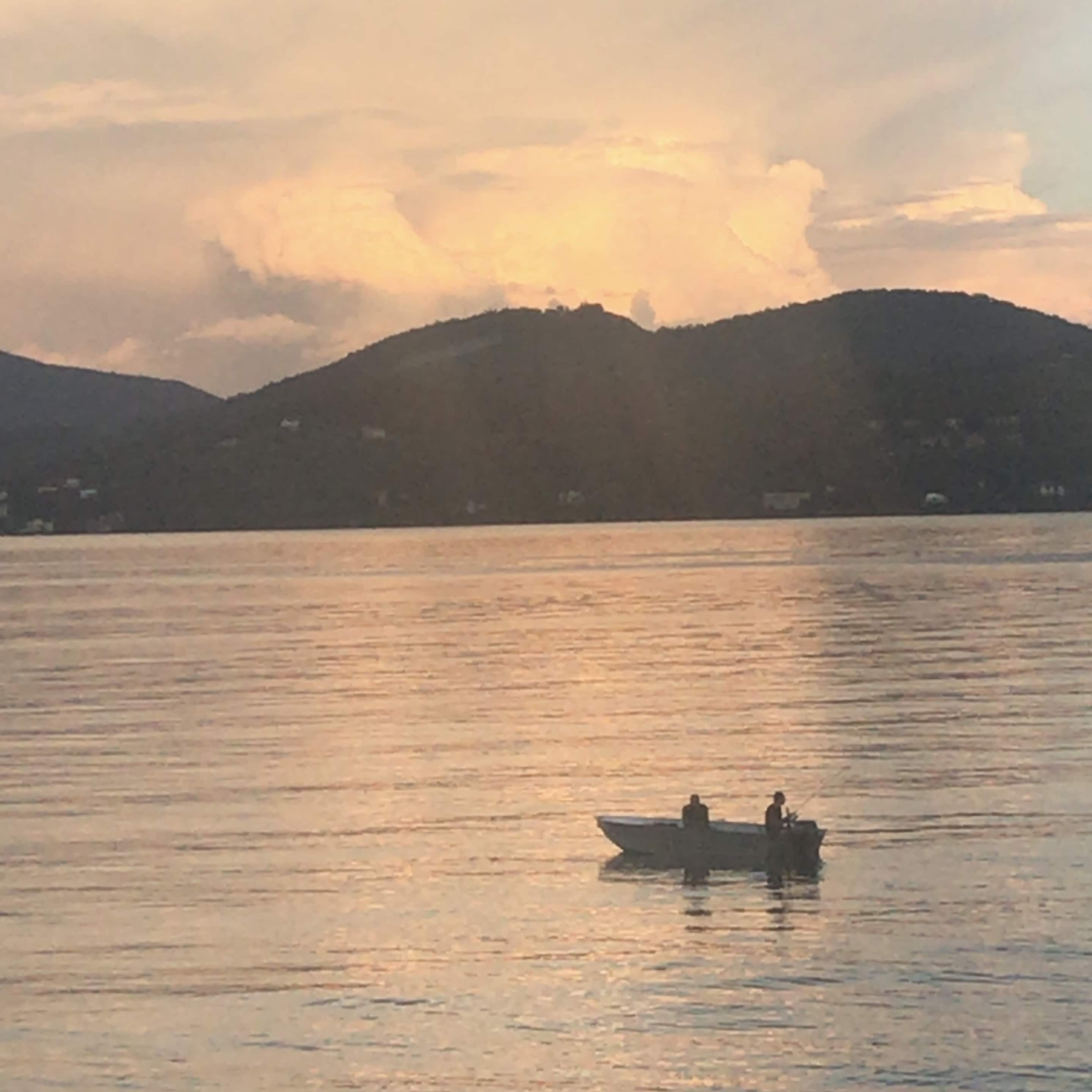 A calm lake with a boat carrying two people, surrounded by hills and a few buildings in the distance.
