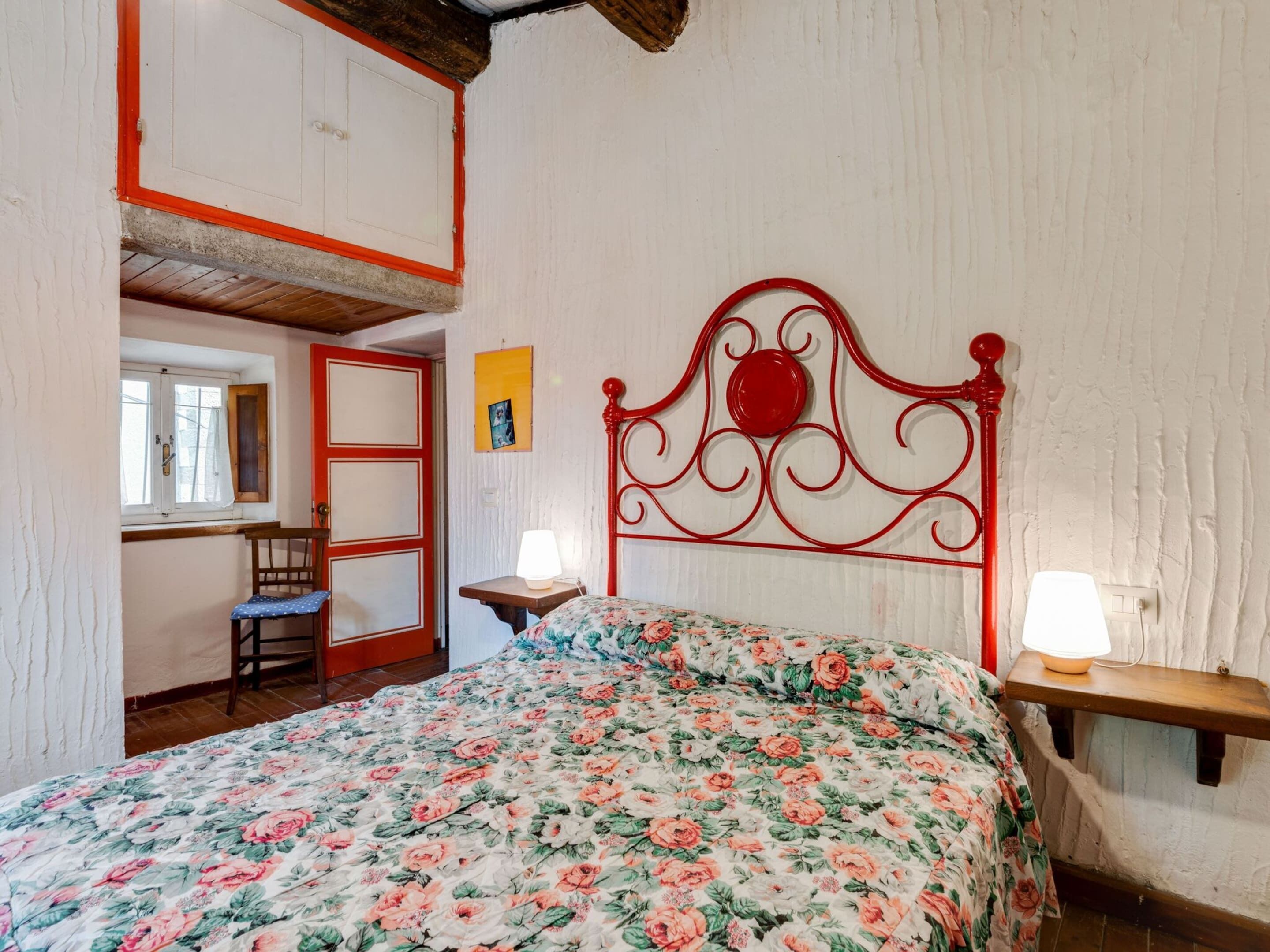 Quarto com edredom estampado, cabeceira de metal vermelha, mesa de cabeceira de madeira com uma luminária e janela com cortinas.