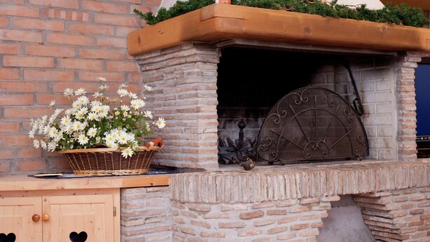 A stone fireplace with a metal door, a wooden bench, and a basket of flowers. - Exquisite Villa in Cesena With Sauna (Cesena)