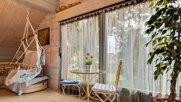 Una habitación con una silla colgante, una mesita redonda pequeña, dos sillas, una planta en maceta y una ventana con cortinas translúcidas.