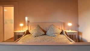 A bed with a patterned bedspread and two pillows, flanked by bedside tables with lamps, in a room with a wooden ceiling and white walls. - Saint-clement Cottage Near the Sea (Osmanville)