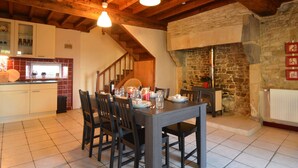 A dining area with a table set for four, wooden beams on the ceiling, a stone fireplace, and a kitchen with white cabinets and a red-tiled backsplash. - Saint-clement Cottage Near the Sea (Osmanville)