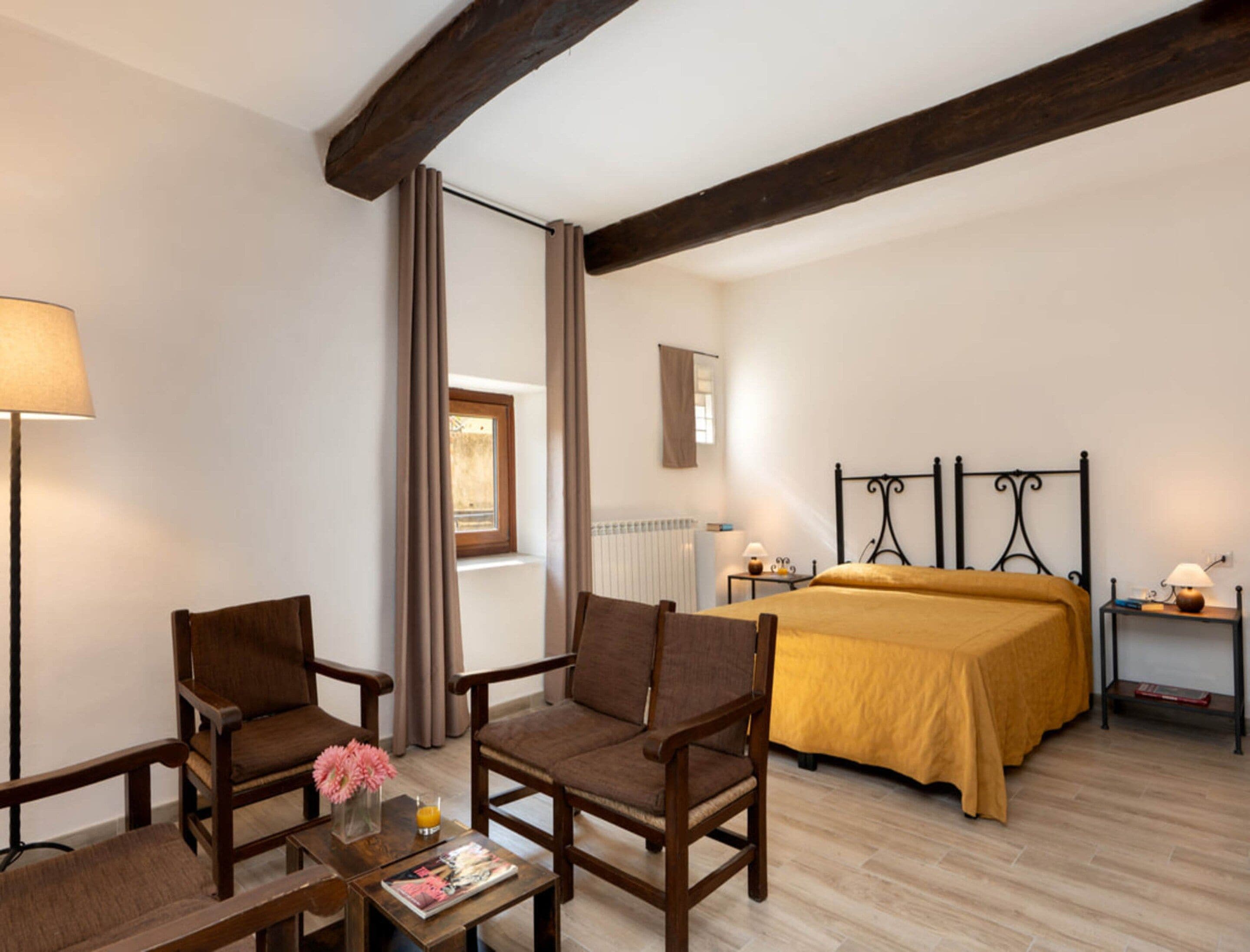 Een hotelkamer met een bed, twee fauteuils, een tafeltje met een tijdschrift en bloemen, een lamp en een raam met gordijnen.