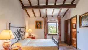 A bedroom with a bed, a nightstand with a lamp, a window with curtains, a painting on the wall, and a door leading to another room. - Villa in Mugello Hills With Private Pool (Vicchio)