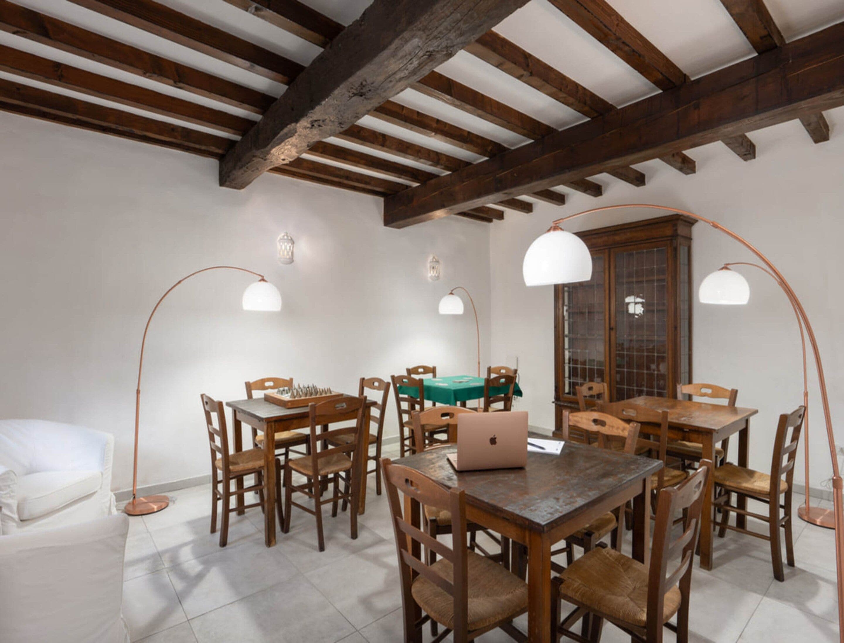 A dining area with wooden tables and chairs, a laptop on one table, and pendant lighting.