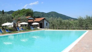 Ein rechteckiger Pool mit klarem, blauem Wasser, umgeben von einer gefliesten Liegefläche. Blaue Liegestühle und weiße Sonnenschirme befinden sich unter Bäumen. Im Hintergrund sind Gebäude mit roten Ziegeldächern und eine hügelige Landschaft zu sehen.