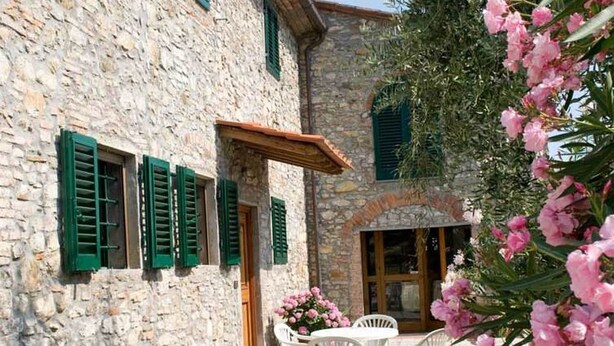 Ein Steinhaus mit grünen Fensterläden, einer Terrasse mit weißen Stühlen und rosa Blumen.