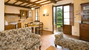 A spacious living room with floral-patterned sofas, a wooden ceiling with exposed beams, a dining area with a table and chairs, and a view of a garden through a glass door. - Rustic Retreat With Pool (Lucignano)