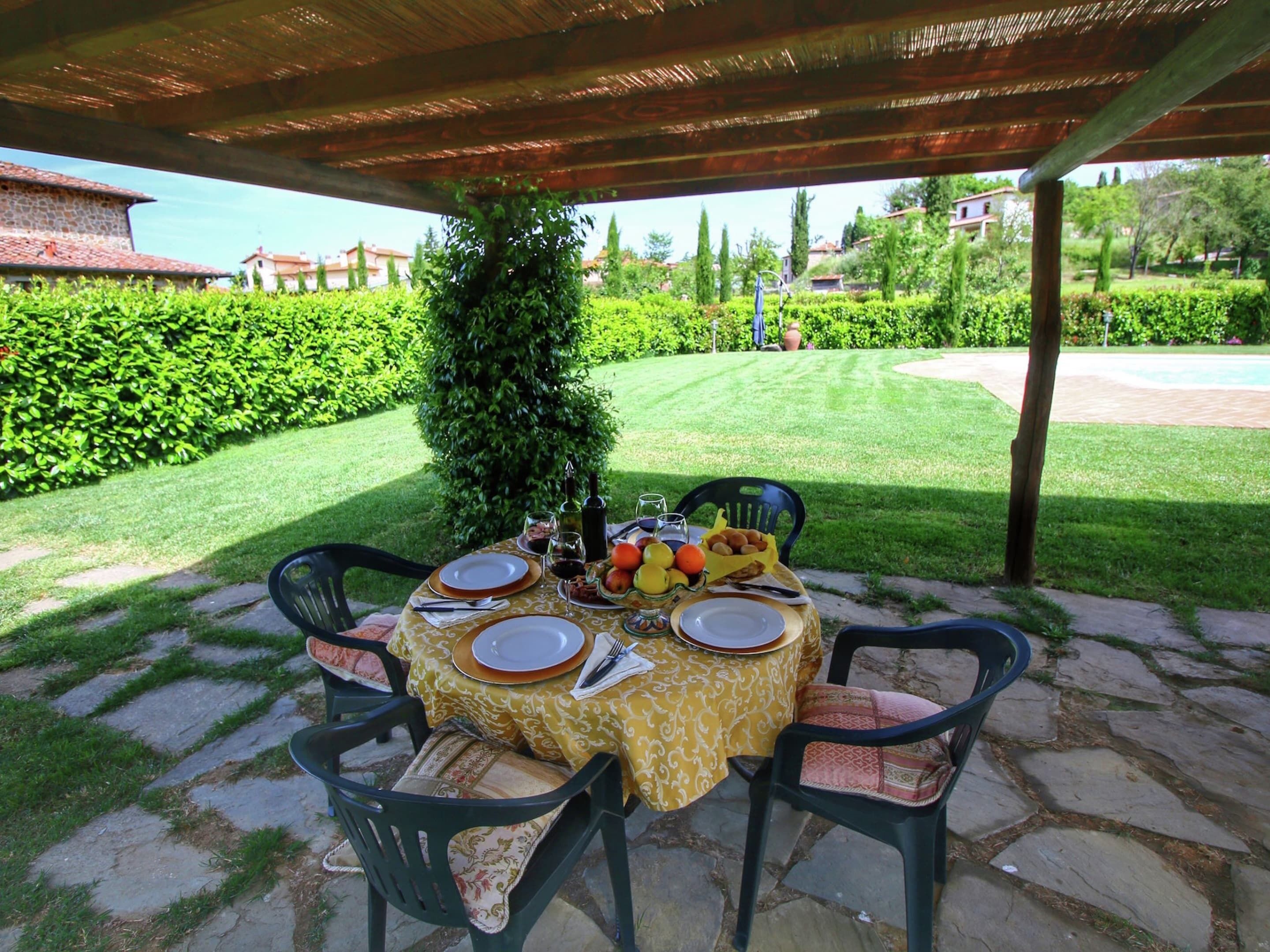 An outdoor dining area with a table set for four, surrounded by stone flooring and covered by a wooden pergola.