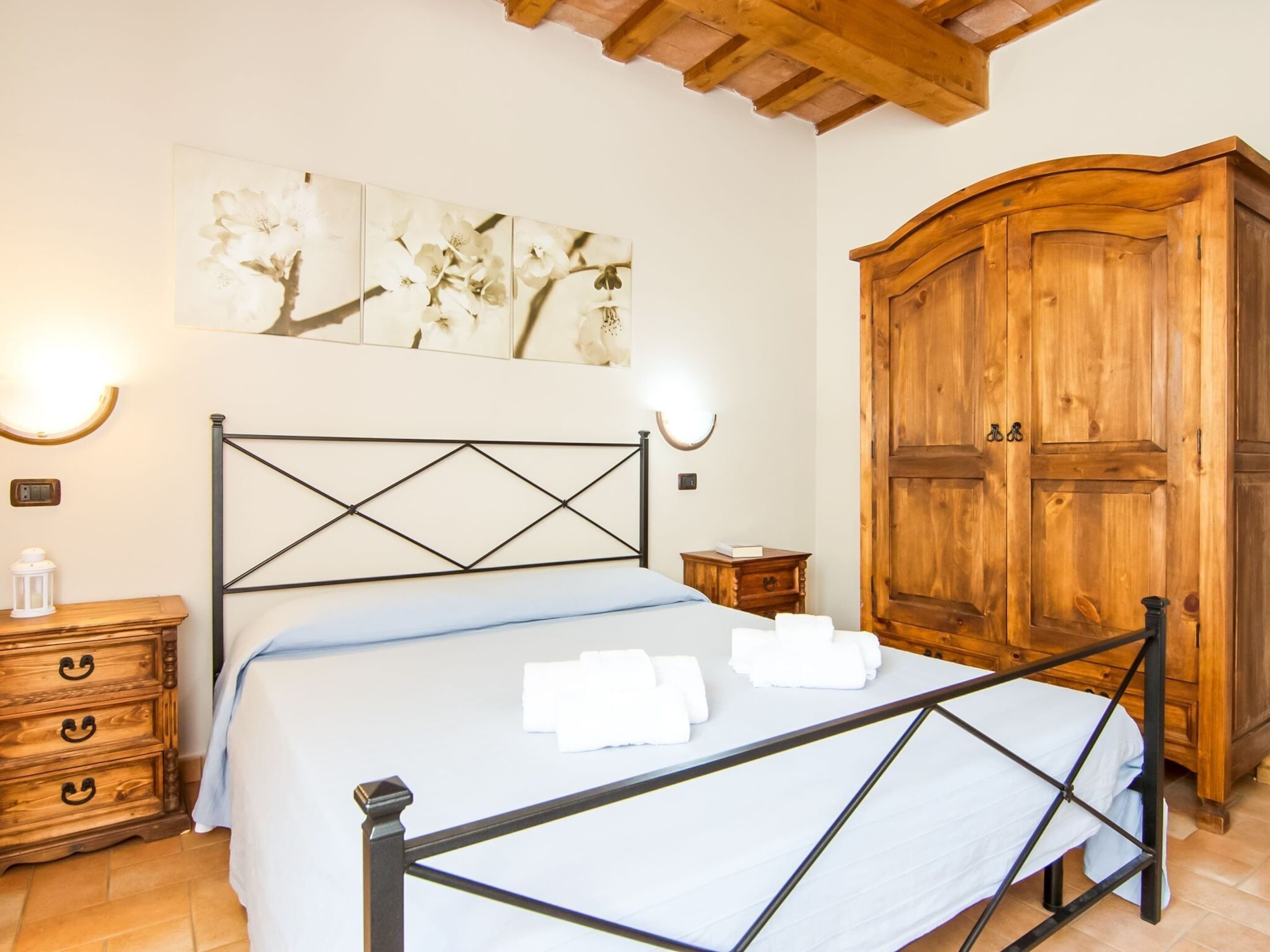 A bedroom with a metal-framed bed, a wooden wardrobe, a nightstand with a lamp, and a wall-mounted artwork.