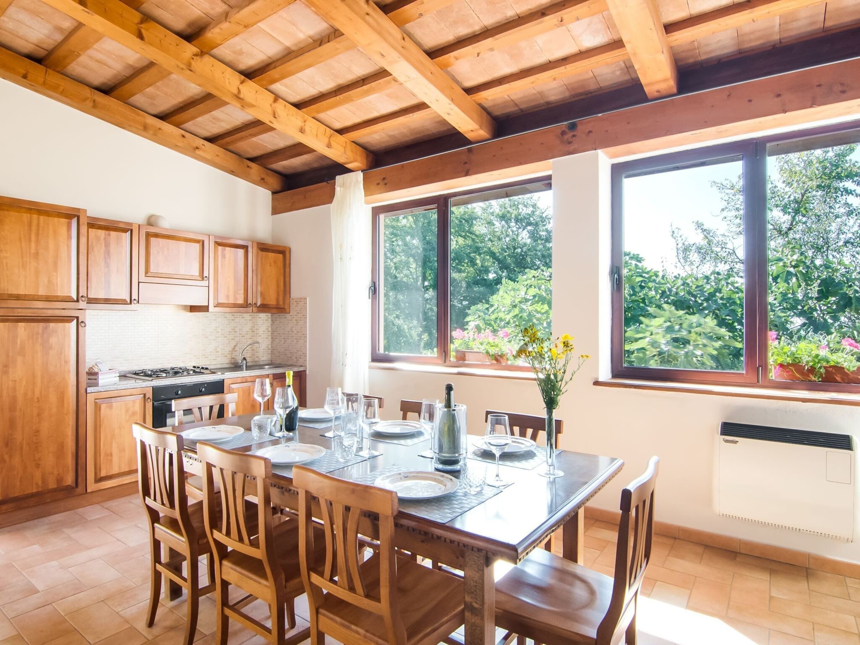 Une cuisine avec des meubles en bois, une table à manger dressée pour quatre personnes et de grandes fenêtres donnant sur un paysage verdoyant.
