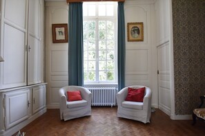 A room with two white armchairs, a radiator, a window with blue curtains, and a patterned wallpaper on the right wall. - 18th-century Normandy Stay (Valognes)