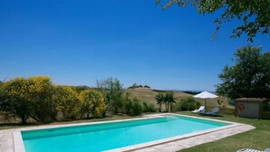 A rectangular swimming pool with a clear blue water, surrounded by a stone border. There are two white lounge chairs and a small white shed with a red lifebuoy. - Belvilla by OYO Fienile (Castelnuovo Berardenga)
