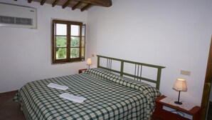 A bedroom with a bed, a window, a bedside table with a lamp, and a wall-mounted air conditioner. - Belvilla by OYO Fienile (Castelnuovo Berardenga)