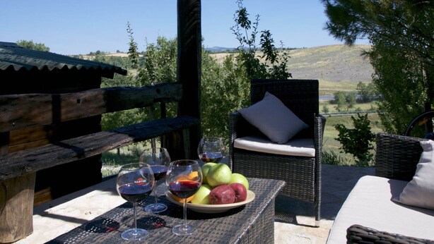 A wooden deck with a wicker chair and a table set with wine glasses and a fruit plate. - Belvilla by OYO Fienile (Castelnuovo Berardenga)