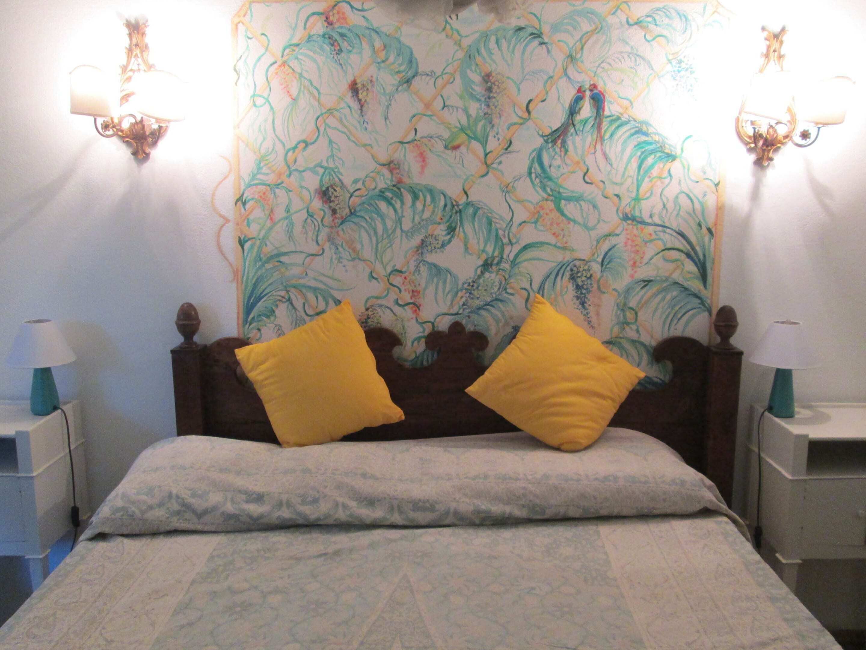 A bed with two yellow pillows, a patterned wall tapestry, and two wall-mounted lamps.