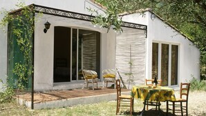 Um edifício branco com alpendre coberto, cadeiras de vime, uma mesa com toalha amarela e uma garrafa.