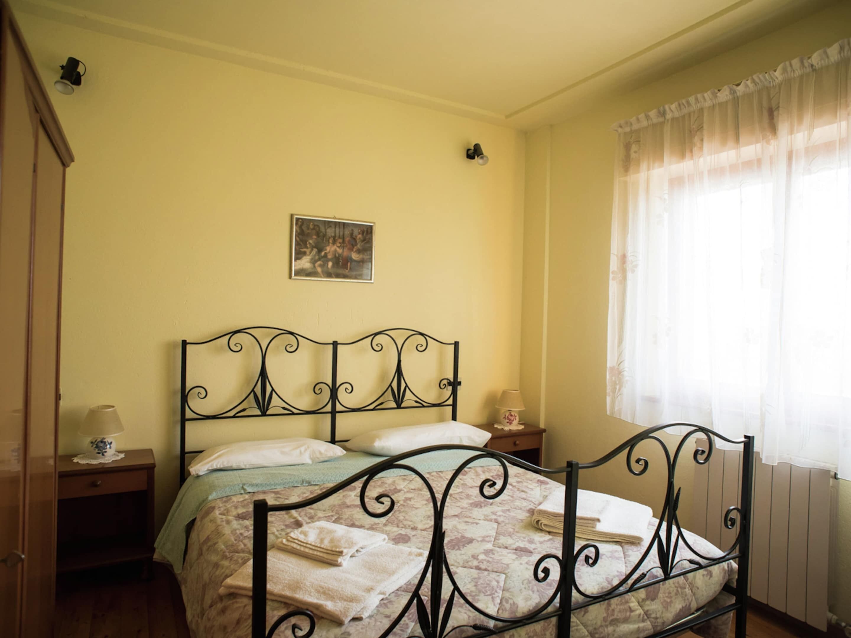 Un dormitorio con cama, mesita de noche, lámpara, un cuadro en la pared y ventana con cortinas.