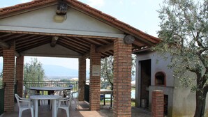 Eine überdachte Terrasse im Freien mit Tisch und Stühlen, einem Ziegelkamin und Blick auf die Landschaft.
