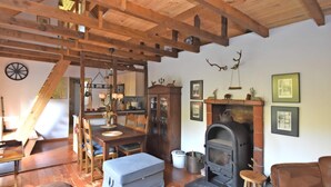 Un interior rústico con vigas de madera a la vista, una zona de comedor con mesa y sillas, una chimenea y un espacio en entrepiso con una cama.