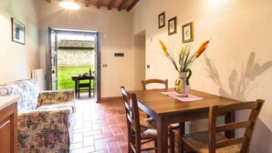 A dining area with a wooden table, chairs, and a floral sofa. There is a view of a garden through an open door.
