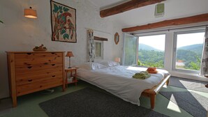 Un dormitorio con cama, cómoda de madera, mesita de noche, lámpara y vistas a la vegetación a través de una ventana.