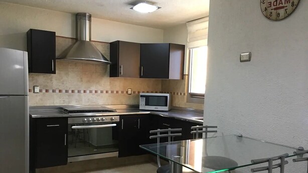 Cocina compacta con estufa a gas, horno, microondas y campana extractora de acero inoxidable. Hay una mesa de vidrio con dos sillas.