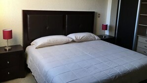Una cama bien hecha con un cabecero oscuro, dos mesitas de noche con lámparas rojas y un armario a la derecha.