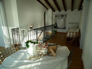 Una habitación con cama, una mesa con un florero y un libro, una ventana con cortinas y una escalera.