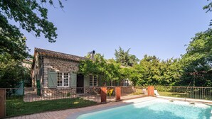 A swimming pool with a brick deck, a stone building with a tiled roof, and a fenced area with greenery.
