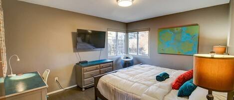 A bedroom with a bed, bedside table, desk, chair, lamp, TV, and artwork.