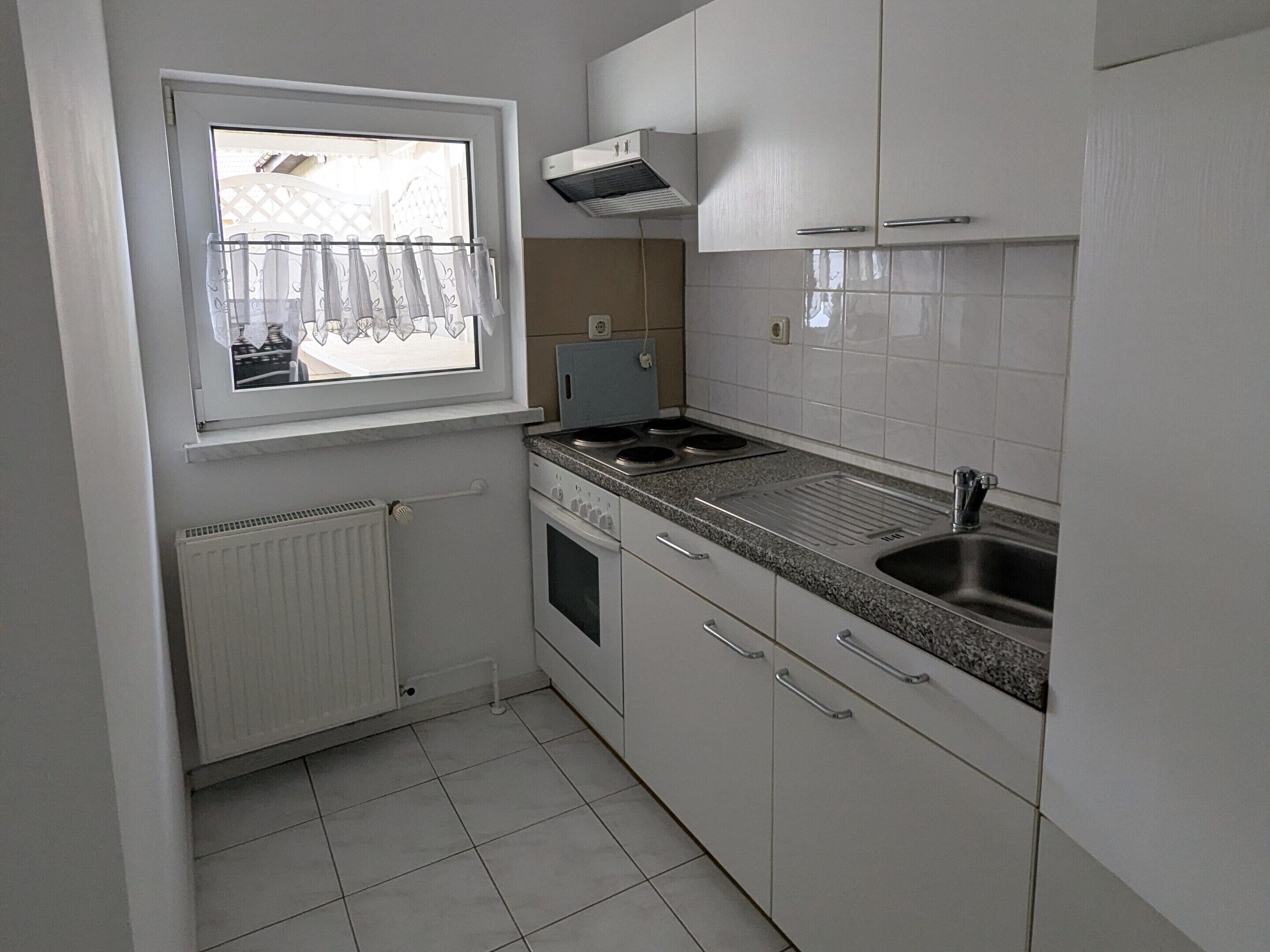 Una cocina pequeña con armarios blancos, una ventana con cortina, una estufa empotrada y un fregadero.