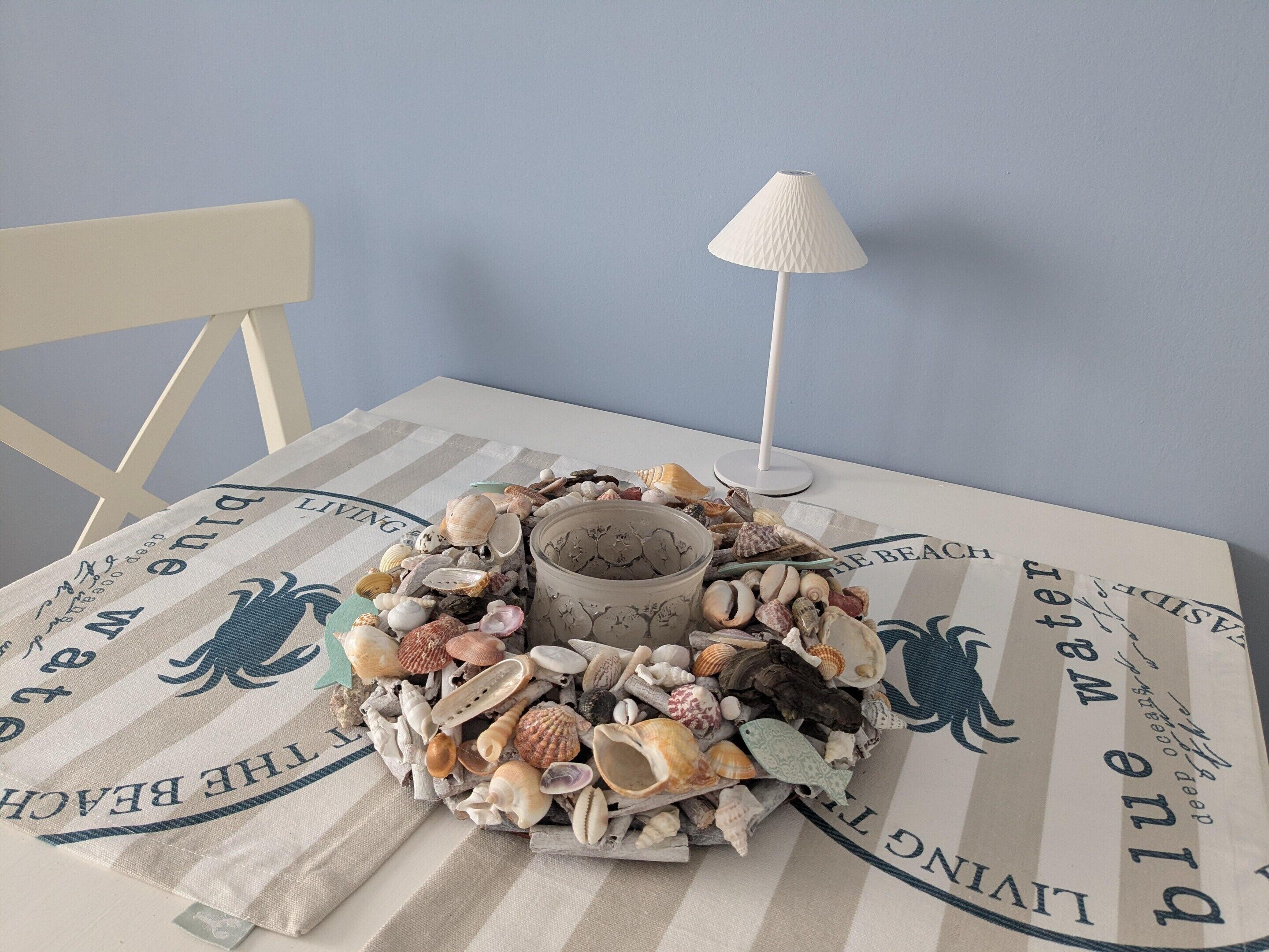 Une table recouverte d’une nappe à rayures, une lampe blanche et un centre de table composé de différents coquillages.