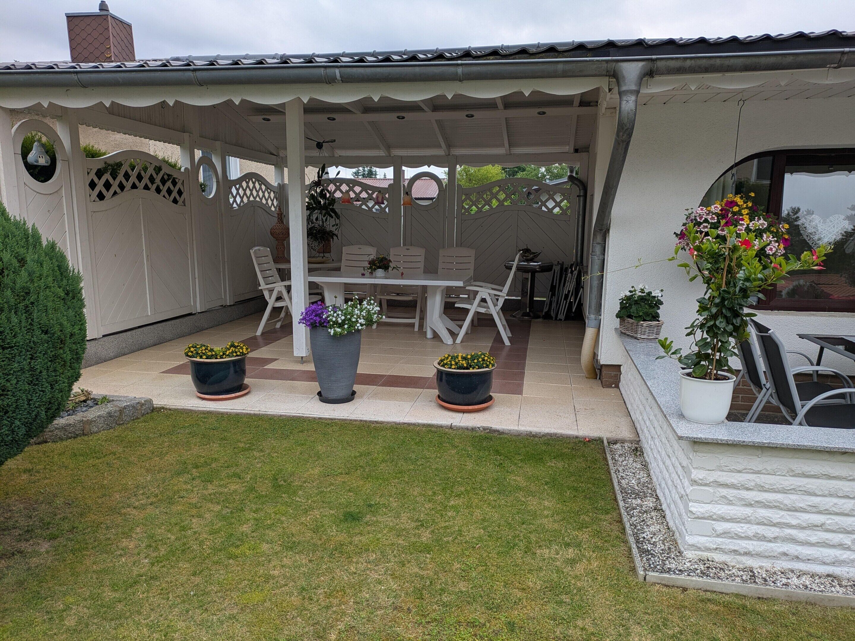 A covered patio area with a table and chairs, potted plants, and a white fence.