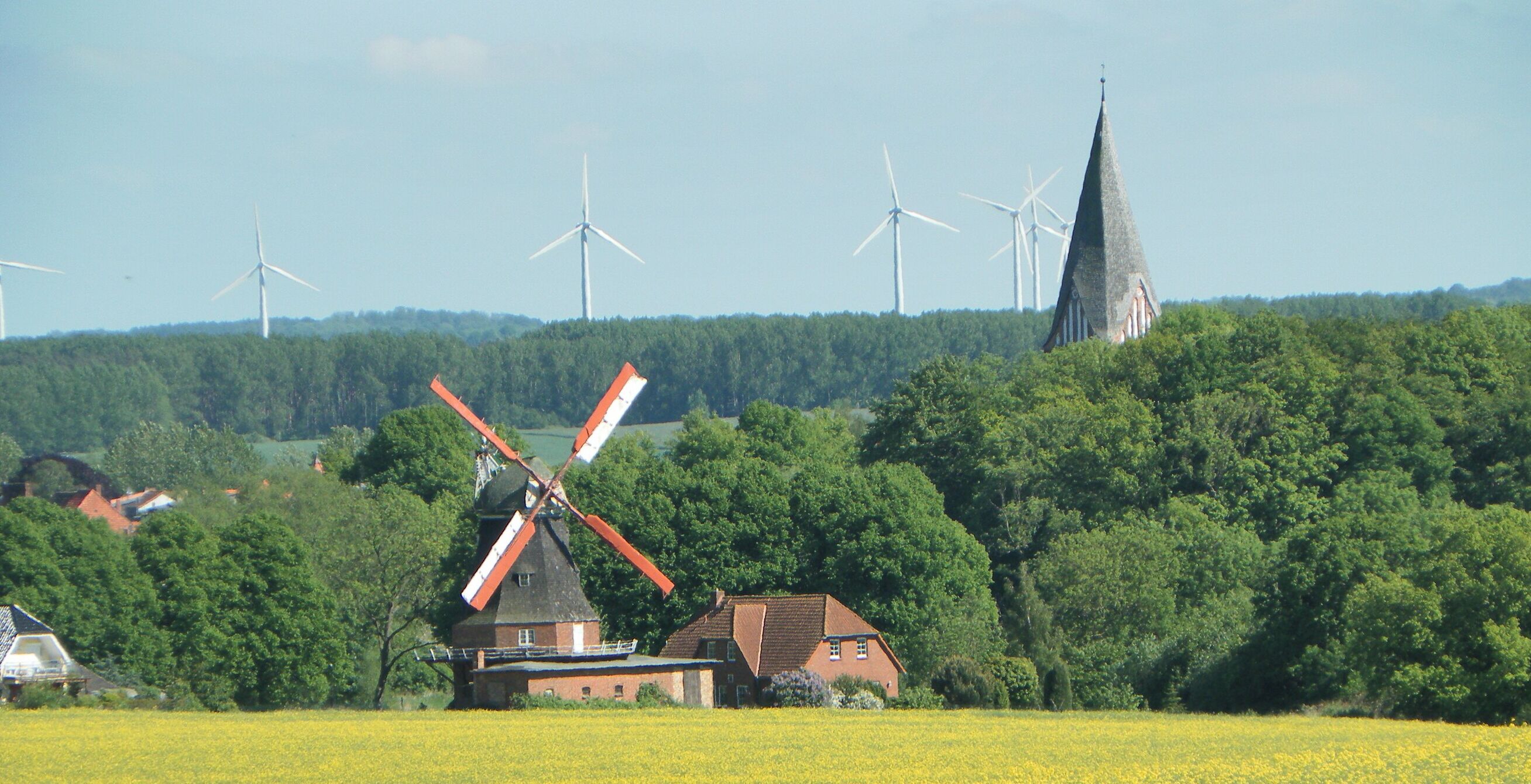 A rural landscape with a traditional windmill, a church spire, and wind turbines in the background.