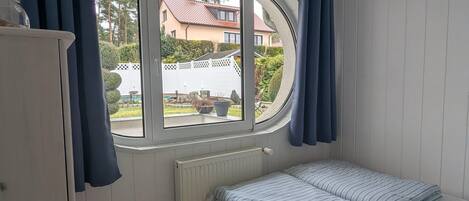 Une chambre avec un lit simple, une fenêtre avec des rideaux, un radiateur et une vue sur une maison et un jardin.